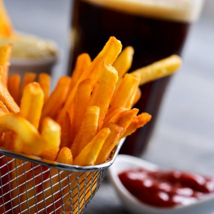 closeup of some appetizing french fries served in a metal basket, a glass with soda and some bowls with ketchup and mayonnaise, on a gray rustic wooden table